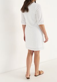 Vit klänning med korta puffärmar, med ögonletsmönster och en något utsvängd nederdel. Buren med bruna sandaler, vilket visar en avslappnad passform.