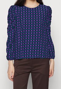 Blusa a scacchi viola e verde con maniche a palloncino e scollatura rotonda, realizzata in tessuto leggero, abbinata a pantaloni marroni.