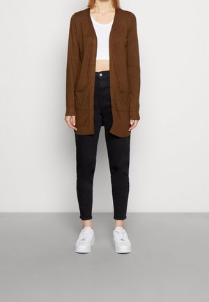 Žena v hnědém kardiganu se zavřeným předkem přes bílý krátký top, černé kalhoty s vysokým pasem a bílé tenisky stojí na šedé podlaze.