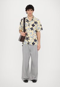 Short-sleeve shirt with yellow base and black and blue floral pattern, paired with light gray wide-leg trousers and a brown shoulder bag.