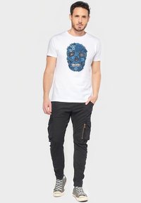 Camiseta blanca con gráfico de calavera floral azul; pantalones cargo negros con bolsillos con cremallera; zapatillas altas grises. Atuendo sencillo y casual.