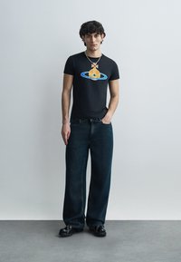 Camiseta negra de manga corta con un diseño gráfico colorido de una nave espacial y Saturno. Combinada con jeans de mezclilla oscuros y de corte holgado, y zapatos negros.