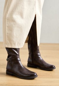 Braune Leder-Stiefel bis zum Knie mit glatter Textur, seitlichem Reißverschluss und Knöchelriemen, kombiniert mit hellen Hosen.