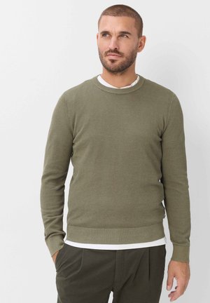 Mann mit kurzen Haaren und Bart steht vor einer weißen Wand, trägt einen olivgrünen strukturierten Pullover über einem weißen Hemd und dunkelolivfarbene Hose.