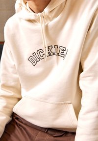 Sudadera color crema hecha de algodón, con un bolsillo delantero y capucha con cordón. Logo "DICKIES" bordado en negro en el pecho.