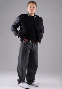 Schwarze Collegejacke mit Lederärmel, gestreiftem Kragen, getragen über einem schwarzen Hemd. Dunkelgraue weite Jeans und weiße Sneakers.