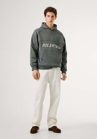 Felpa grigia con cappuccio e tasca a marsupio, con testo "PJL1973Club", abbinata a jeans bianchi e scarpe slip-on marroni. Design semplice e casual.