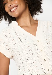 Balta marškiniai su trumpomis rankovėmis, pasižymintys tekstūruota medžiaga su akytomis skylutėmis ir metaliniais mygtukais priekyje.