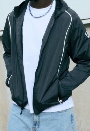 Personne portant une veste noire zippée avec un liseré blanc, une chemise blanche, un jean bleu clair et un collier chaîne argenté, mains dans les poches de la veste.