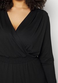 Femme portant une robe portefeuille noire à manches longues avec une taille élastique, cheveux bouclés partiellement visibles, sur un fond clair uni.