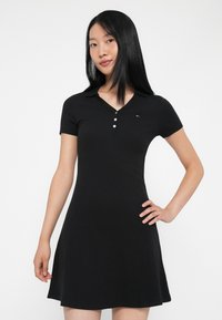 Juodas polo suknelė, pagaminta iš įlinkto mezginio audinio, turi suknelė su mygtukai ant kaklo ir trumpomis rankovėmis. Ant krūtinės matomas mažas logotipas.