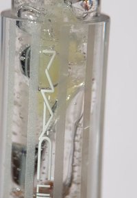 Tube en verre transparent, présentant des rayures verticales en verre dépoli, contenant un liquide jaune avec des bulles, et arborant un logo blanc sur le côté.