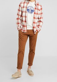 Rood-witte geruite shirt over een wit graphic t-shirt, gecombineerd met een bruine chino en beige schoenen. Casual outfit met opgerolde broekspijpen.