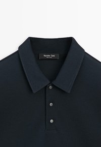 Dunkelblaues Poloshirt mit drei metallischen Knöpfen und einem Etikett mit der Aufschrift "Massimo Dutti Made in China" im Krageninneren.