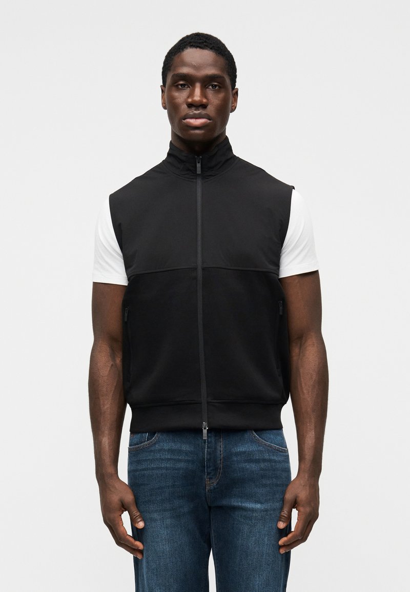 Μαύρο αμάνικο γιλέκο με φερμουάρ και ψηλό γιακά, φορεμένο πάνω από λευκό μπλουζάκι, σε συνδυασμό με μπλε τζιν τζιν, με κομψό, μίνιμαλ σχεδιασμό.