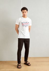 White T-shirt with red and blue graphic text, paired with black slim-fit pants and black sandals with adjustable straps. Plain background.