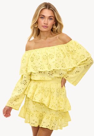 Femme portant une robe jaune à épaules dénudées en tissu ajouré avec des volants, debout devant un fond blanc uni, avec des boucles d'oreilles florales dorées.