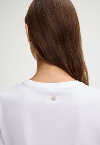 Wit katoenen T-shirt met een ronde hals en een klein metalen logodetail aan de achterkant. Soepele textuur, ontspannen pasvorm.