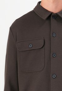 Camisa de botones marrón oscuro hecha de tela texturizada, con un bolsillo en el pecho y botones negros en contraste. Diseño con cuello y un corte ajustado.