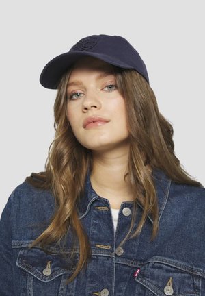 Giovane donna con lunghi capelli castani che indossa un cappellino da baseball scuro e una giacca di jeans blu su uno sfondo chiaro uniforme.