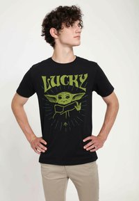 Sort t-shirt med et grafisk motiv af en karakter med store ører, som holder to fingre op. Teksten "LUCKY" står i grønt over billedet.