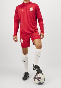 Rød fodbolduniform med langærmet overdel og shorts. Har gul logo og detaljer, parret med hvide sokker og sorte fodboldstøvler. Sort og hvid fodbold.
