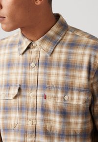 Camisa de botão xadrez nas cores bege, azul e creme. Apresenta dois bolsos no peito, fechos de botão e uma etiqueta com o logotipo da Levi's na parte da frente.