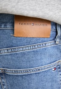Denim jeans in light blue with a brown leather label displaying "TOMMY HILFIGER." Features fading and stitching details on pockets.