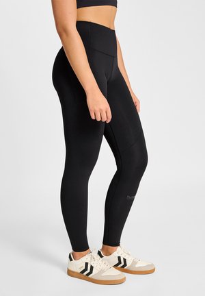 Femme portant un legging noir taille haute et des baskets blanches, debout devant un fond clair uni.