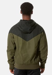 Olivgrüne Jacke mit einem oberen Abschnitt im schwarzen Chevron-Muster; leichtes Material, mit Kapuze, elastischen Bündchen und Saumdetail.