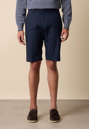 Uomo che indossa pantaloncini sartoriali blu navy, camicia a righe a manica lunga e mocassini in suede marrone con nappe, in piedi davanti a uno sfondo beige.