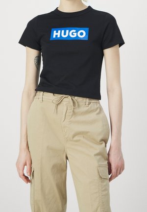 Czarny bawełniany t-shirt z niebieskim prostokątnym logo "HUGO". Model połączył go z beżowymi spodniami cargo z bocznymi kieszeniami i wiązanym w pasie.