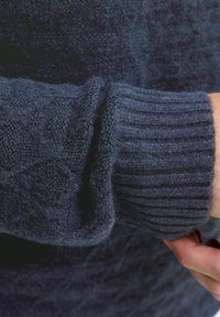 Pogled od blizu na ruku osebe, ki prilagaja manšeto temno modrega teksturiranega pletenega puloverja.