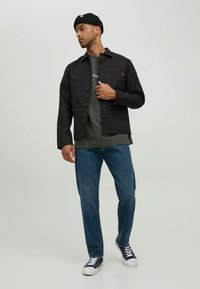 Sort bomuldsjakke med knapper, grøn t-shirt under, blå jeans og marineblå sneakers. Modellen har en sort beanie på.