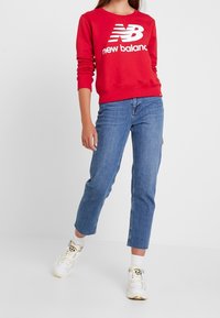 Felpa rossa con logo "New Balance" bianco, abbinata a jeans in denim azzurro chiaro. Il modello indossa sneakers color crema con dettagli neri.