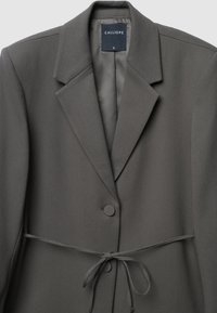 Grauer Blazer mit Reverskragen, Ein-Knopf-Verschluss und Taillengeband. Verfügt über eine glatte Textur und ein Etikett, das die Größe S anzeigt.