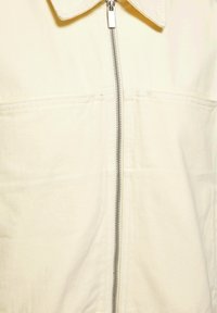 Prim-plan al unei jachete din material crem cu fermoar vertical argintiu și cusături orizontale vizibile sub guler.