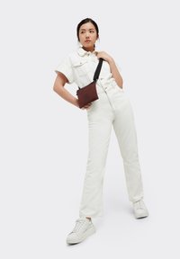 Brun tyg crossbody-väska med svart rem, med ett cirkulärt tryck på framsidan. Modellen bär en vit denimoutfit och sneakers.