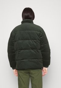 Padded green jacket with a high collar, featuring a soft texture. The back shows baffle stitching and ribbed cuffs, worn over olive green pants.