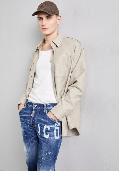 Beige Flanellhemd über einem weißen Tanktop, kombiniert mit blauen, verranzten Jeans, die weißen "DSQUARED2"-Schriftzug und Spritzakzente aufweisen.