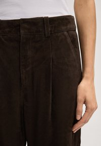 Braune Wildlederhosen mit glatter Textur, die eine maßgeschneiderte Passform und scharfe Falten aufweisen, ergänzt durch einen passenden Gürtelschlaufen.