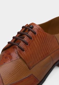 Καφέ δερμάτινο επίσημο παπούτσι με υφασμάτινα πάνελ, σκούρα καφέ κορδόνια και λεπτομερείς ραφές σε λευκό φόντο.