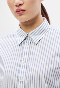 Cămașă cu dungi, cu nasturi, în alb și albastru deschis, cu guler clasic și logo mic brodat, având o textură netedă și material din bumbac.