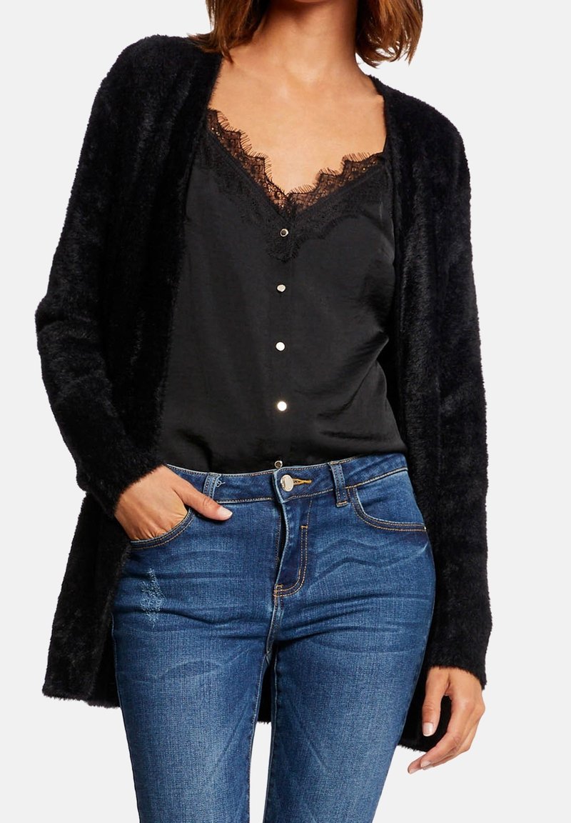 Femme portant un cardigan noir duveteux sur un débardeur noir orné de dentelle, avec un jean bleu, main dans la poche, sur fond blanc.