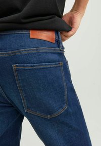 Jeans de mezclilla de un azul oscuro que presentan un parche de cuero en la parte trasera de la cintura y costuras naranjas. El corte incluye bolsillos traseros estándar.
