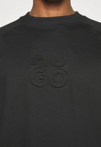 Gros plan d'un sweat-shirt noir avec un lettrage "HUGO" en relief texturé centré sur la poitrine, porté par une personne à la peau foncée.