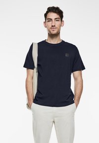 Tmavě modré tričko s kulatým výstřihem a krátkými rukávy, se decentním logem na hrudi. V kombinaci se světlými kalhotami a světlou taškou.