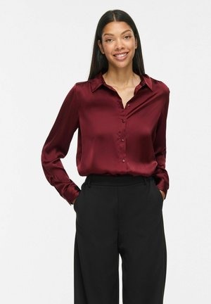Femme souriante aux longs cheveux foncés, portant un chemisier en satin bordeaux boutonné et un pantalon noir taille haute, les mains dans les poches.