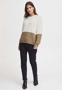 Strukturierter Strickpullover in Weiß und Braun mit einem Kontrastfarbblockdesign, Rundhalsausschnitt und gerippten Bündchen. Kombiniert mit schwarzen Hosen.
