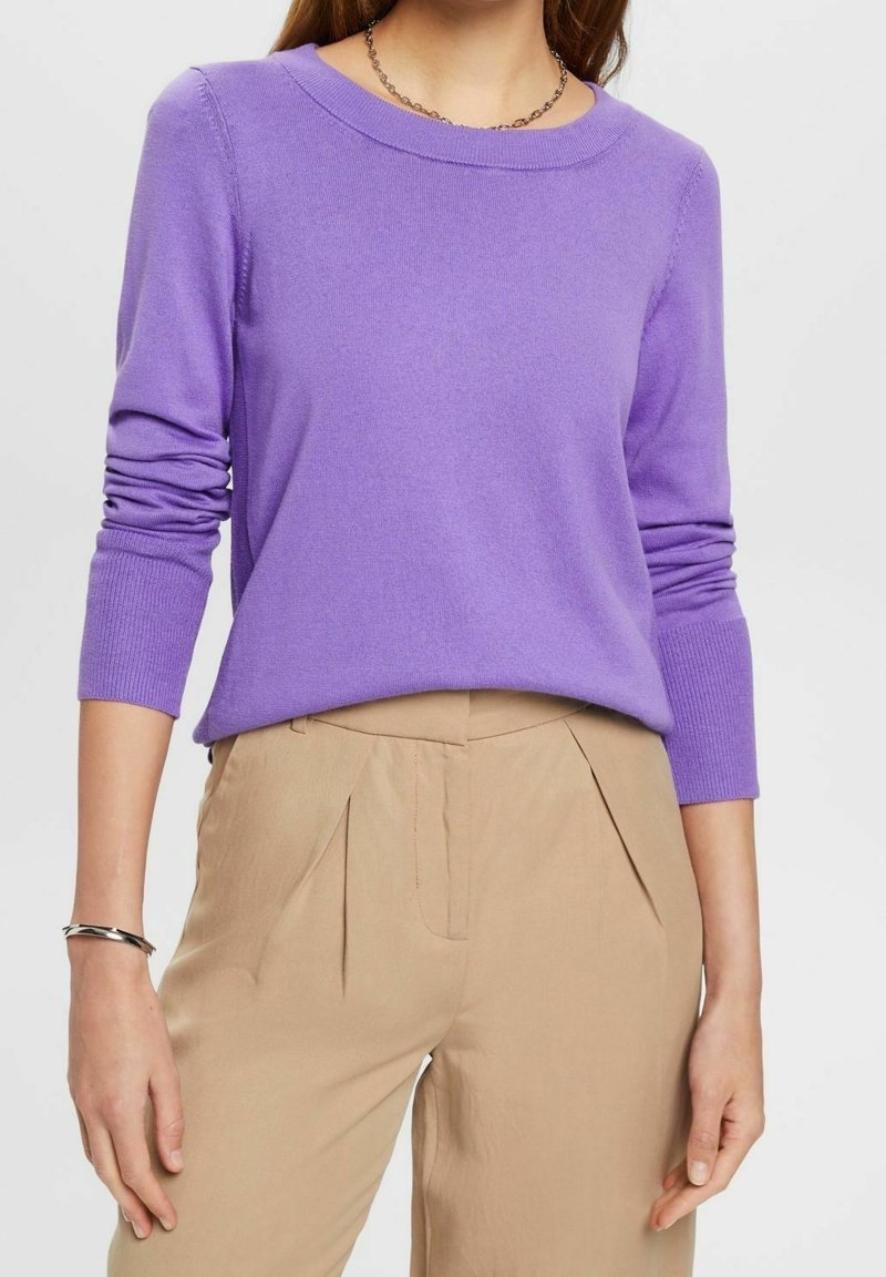 Lila stickad tröja med långa ärmar och rund halsringning, i kombination med beiga veckade byxor; lätt, jämn textur och avslappnad passform.
